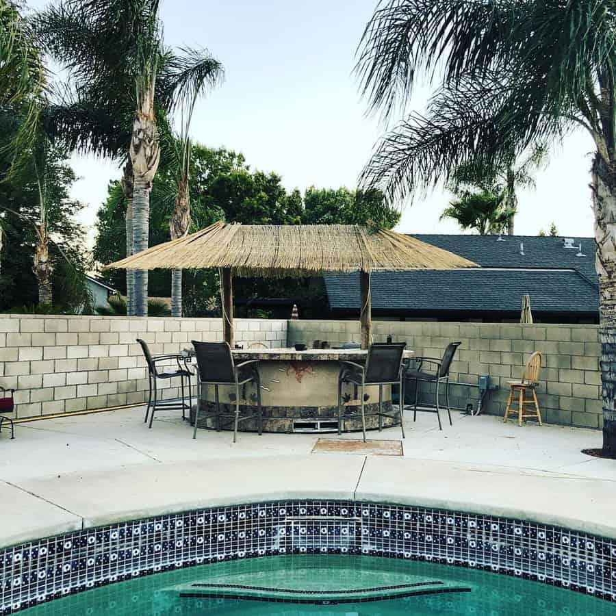 Poolside view with a tiki-style bar, chairs, and palm trees in a backyard setting