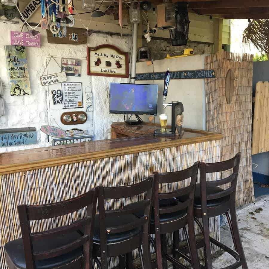 Outdoor tiki bar with four wooden stools, bamboo decor, wall signs, knick-knacks, and a TV screen showing a soccer game