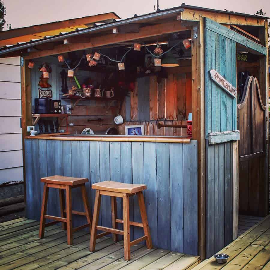 A small outdoor wooden bar with two stools, decorated with hanging lights and signs, on a wooden deck