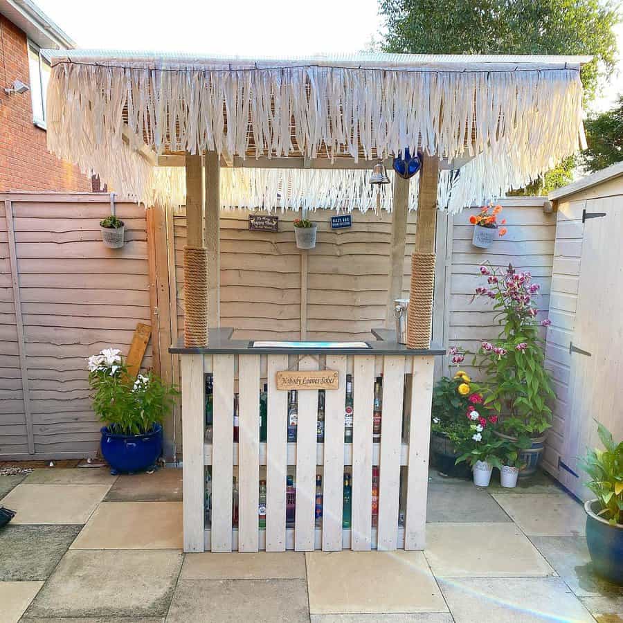 A tiki bar with a thatched roof stands in a patio, potted plants surround the area, and a wooden fence serves as the backdrop