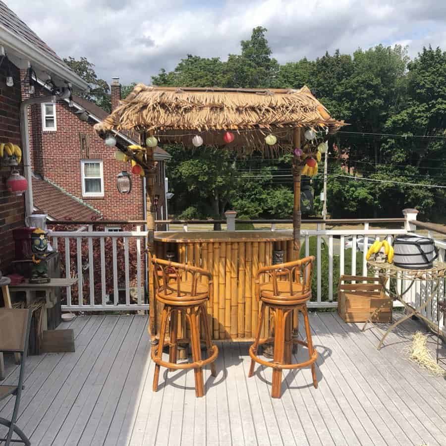 Tiki bar with two stools on a wooden deck, surrounded by greenery, houses, and colorful decorations under a cloudy sky