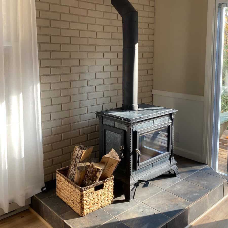 Rustic wood stove on a tiled hearth, set against a painted brick wall with a woven basket of firewood for a cozy and inviting ambiance.