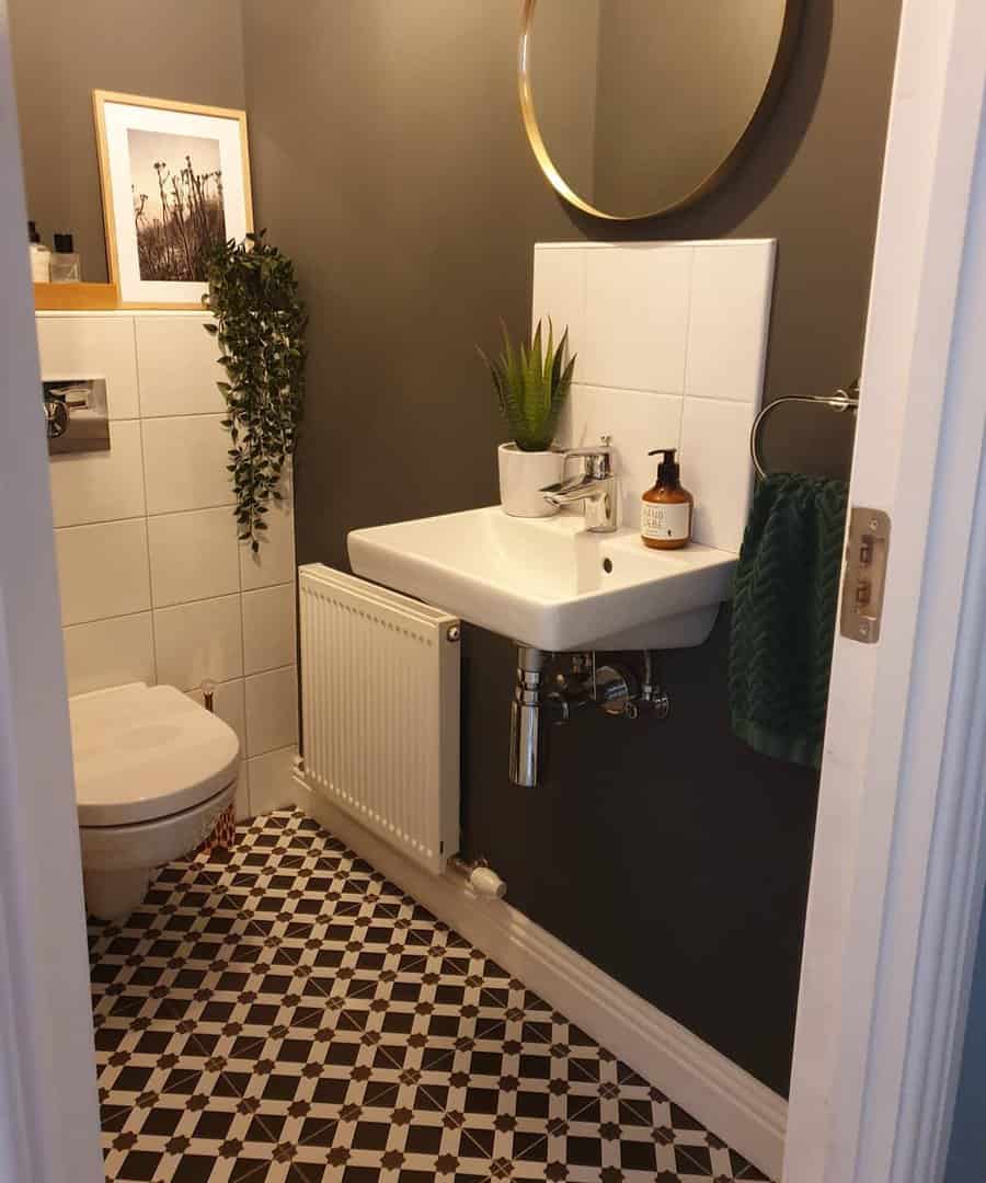 Cozy bathroom with simple white tile backsplash, dark walls, patterned black-and-white floor tiles, and gold round mirror.