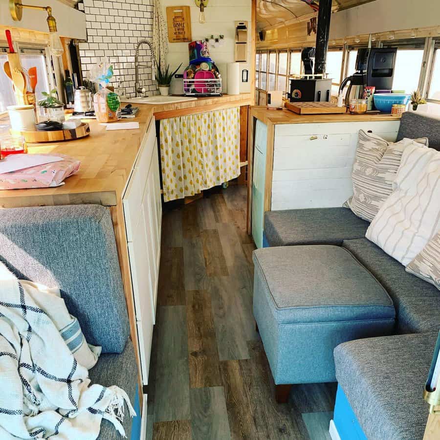 Cozy converted bus interior with kitchen, sofa, and wooden countertops, bright and welcoming with pillows and a striped curtain