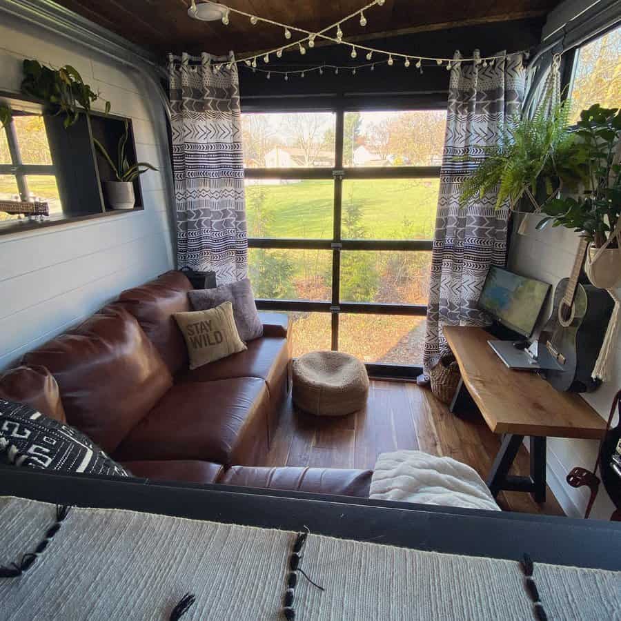 RV interior with a brown leather sofa, desk with a monitor, boho curtains, potted plants, string lights, and outside greenery view