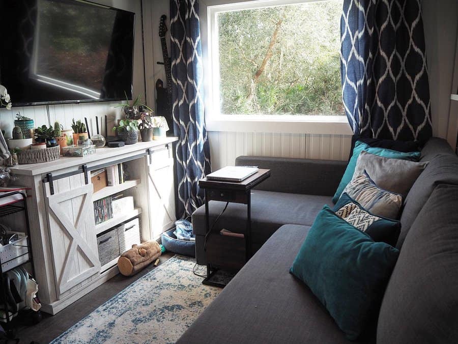 RV living room with a dark gray sectional, blue patterned curtains, a rustic barn-style console, and lush greenery outside the window