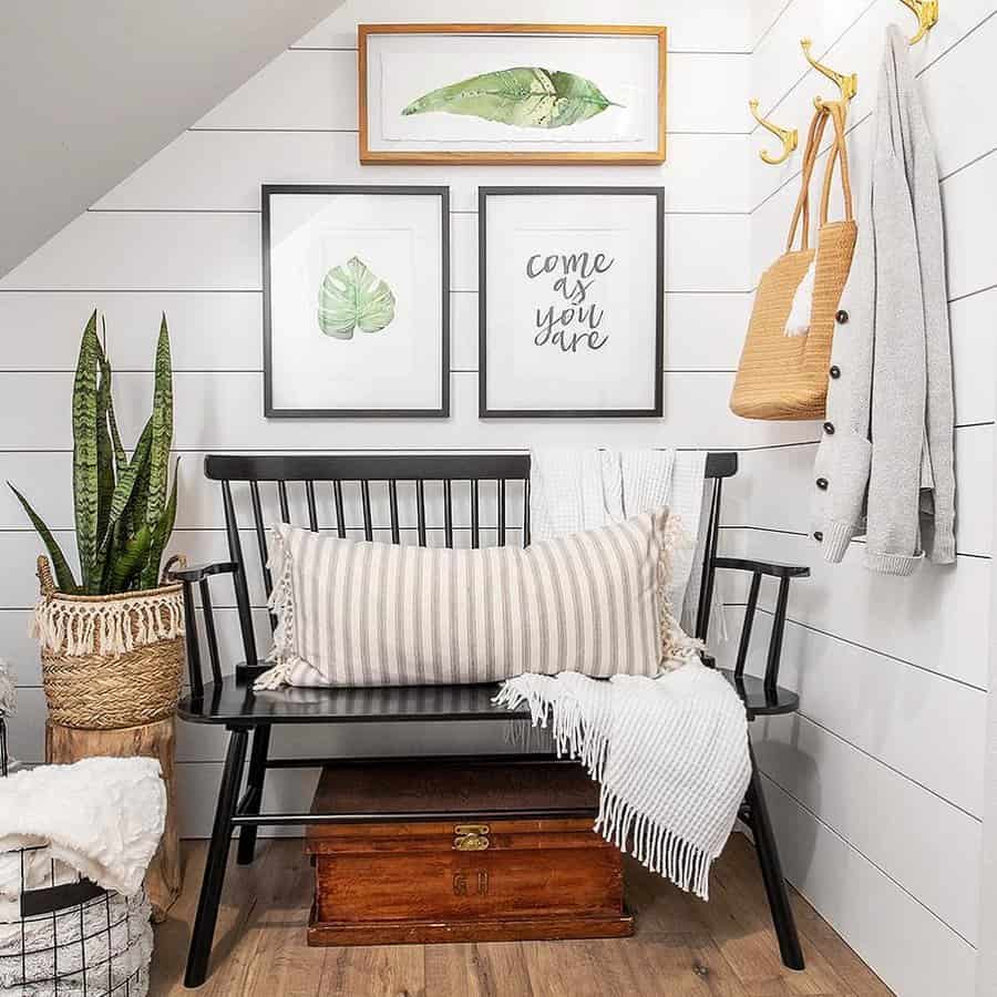 Cozy entryway with a black bench, striped cushion, plants, and wall art featuring leafy framed prints; hooks hold a coat and bag