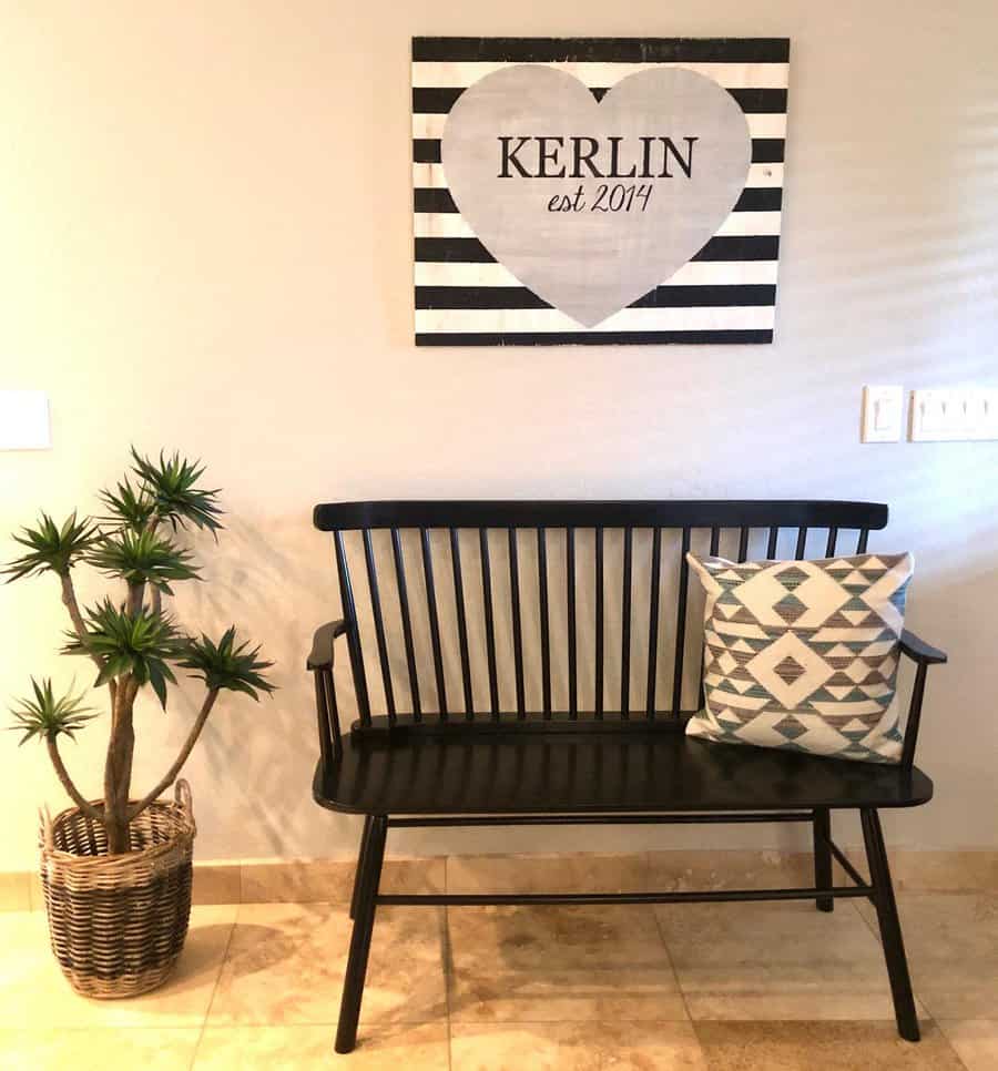 A black bench with a patterned pillow sits below a striped wall sign, and a potted plant is beside the bench on a tiled floor