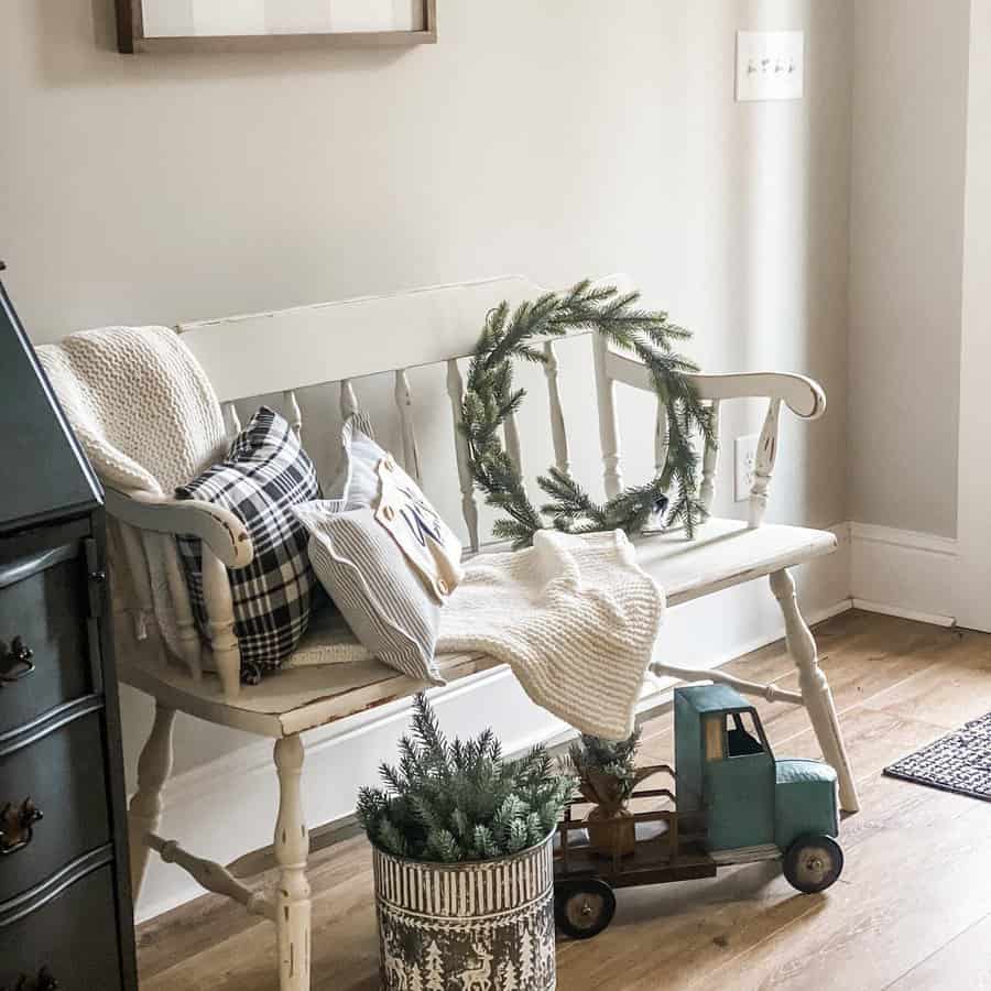 White wooden bench with pillows and a pine wreath, a toy blue truck, and a potted plant on a wood floor in a cozy room