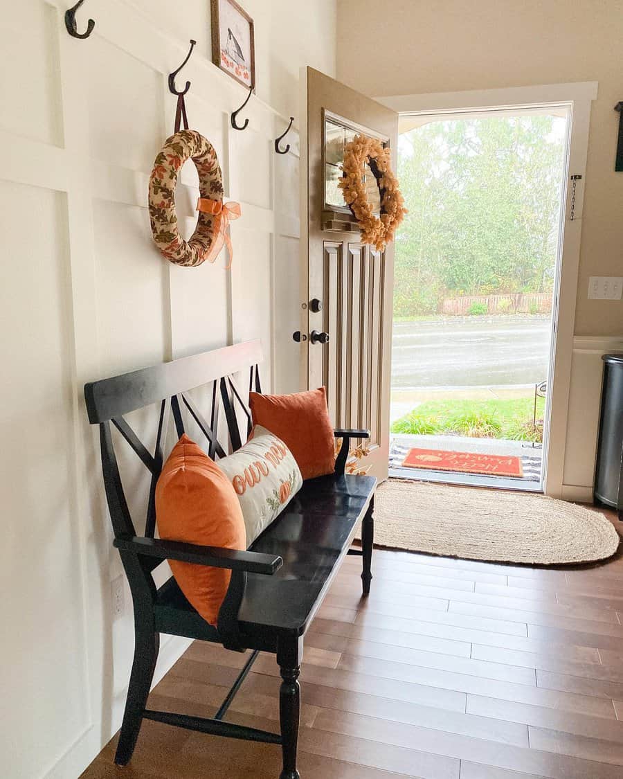 Cozy entryway with a black bench, orange pillows, autumnal wreaths on wall hooks, and an open door showing a rainy street outside