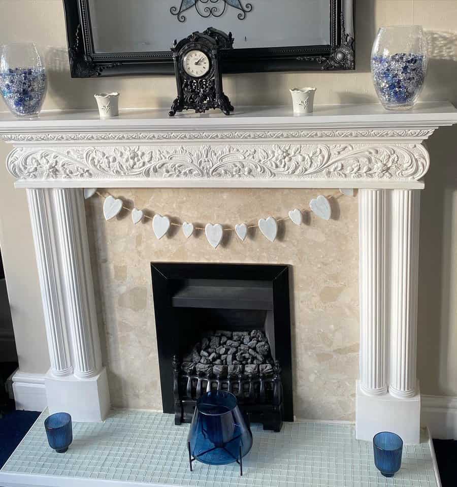 A traditional white fireplace with intricate detailing, decorated with a heart-shaped garland, vintage clock, and blue glass vases, creating an elegant and cozy setting