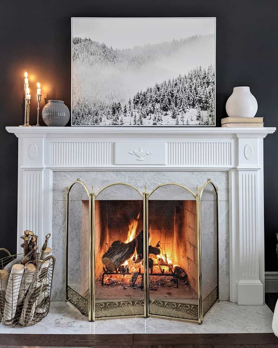 A classic white marble fireplace with a gold screen, decorated with a black and white winter landscape print, candles, and natural wood accents, creating a warm and cozy vibe