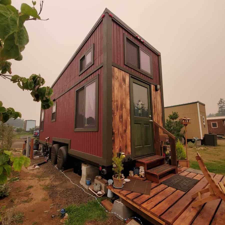 A small brown and red tiny house on wheels with a wooden deck