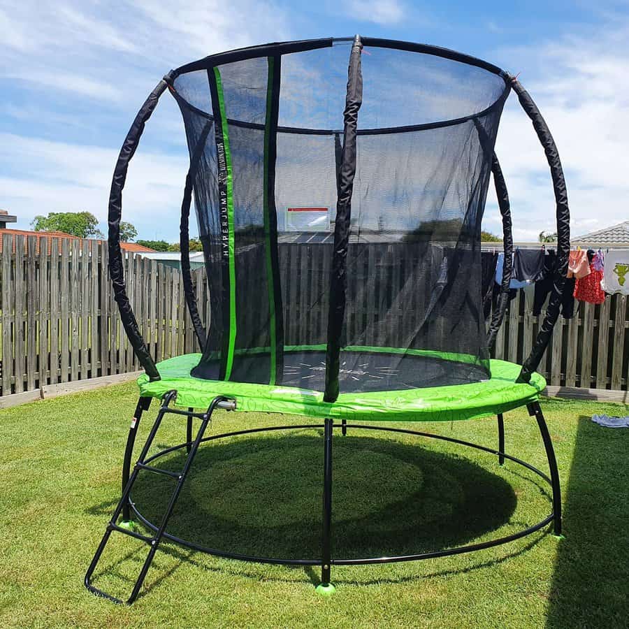 A round trampoline with a black safety net, green padding, and a small ladder stands on grass in a backyard