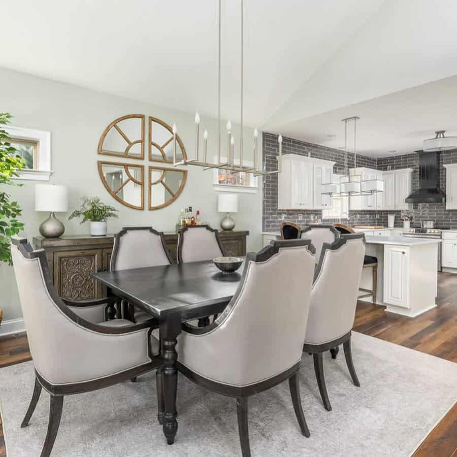 Dining room with a dark table, six cushioned chairs, a large round wall mirror, and a modern chandelier. Open view to the kitchen