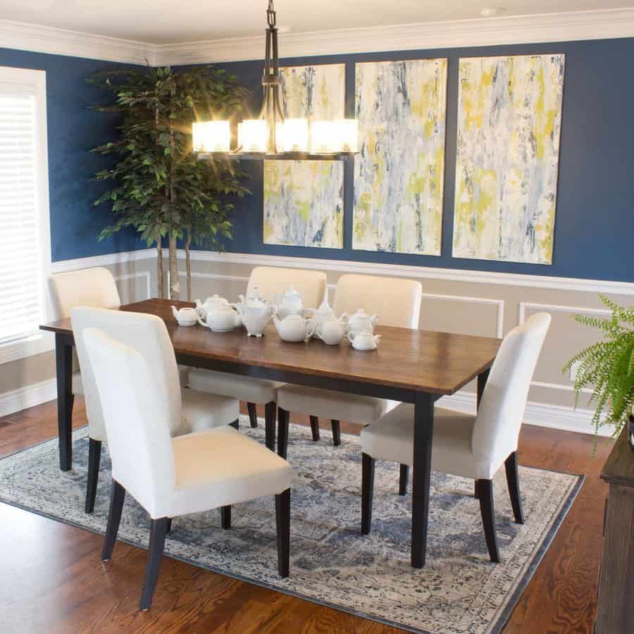 A dining room with a wooden table, six white chairs, a chandelier, abstract artwork on blue walls, and a large green plant