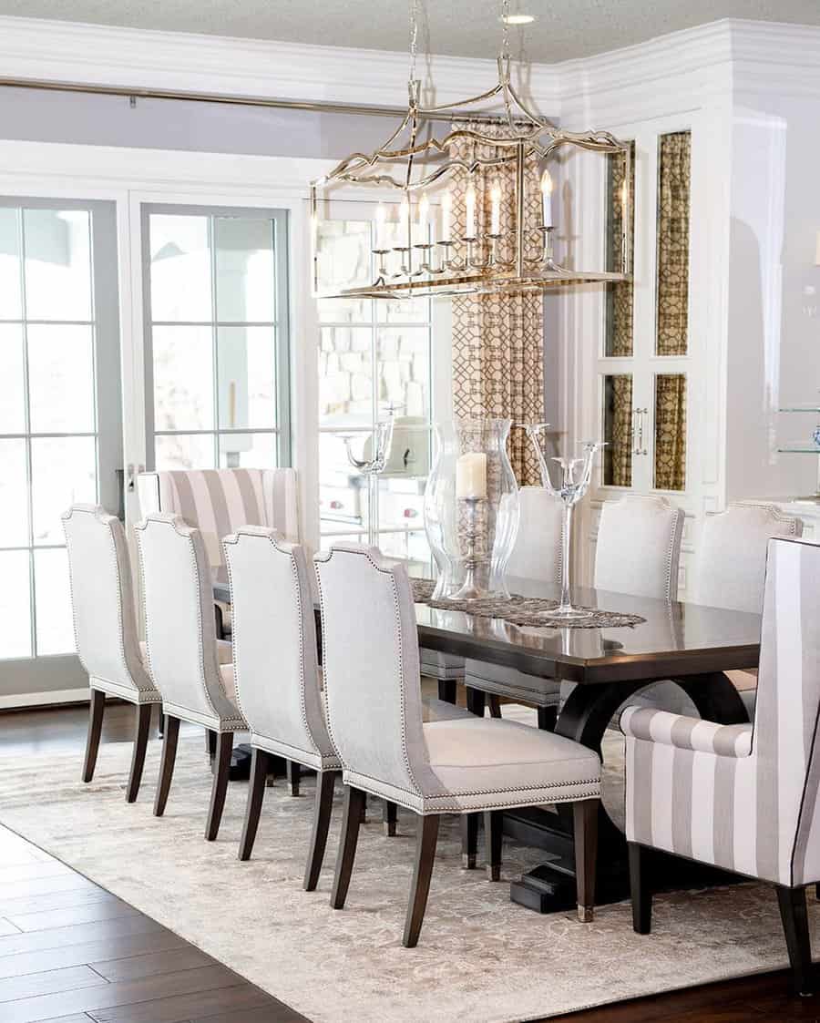 Elegant dining room with a long table, white chairs, striped armchairs, a chandelier, and large windows, featuring a neutral color palette