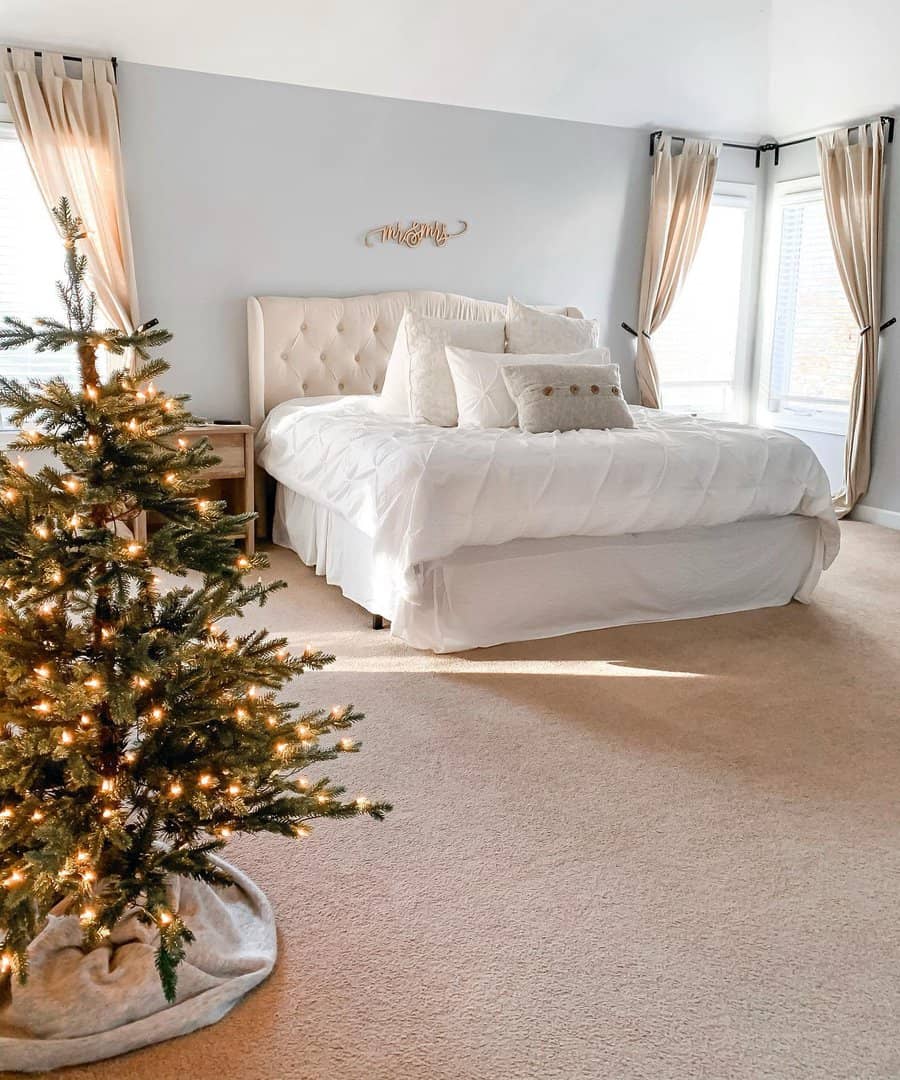 Bedroom with a white bed, beige curtains, and a small Christmas tree with lights in the corner, under soft natural light