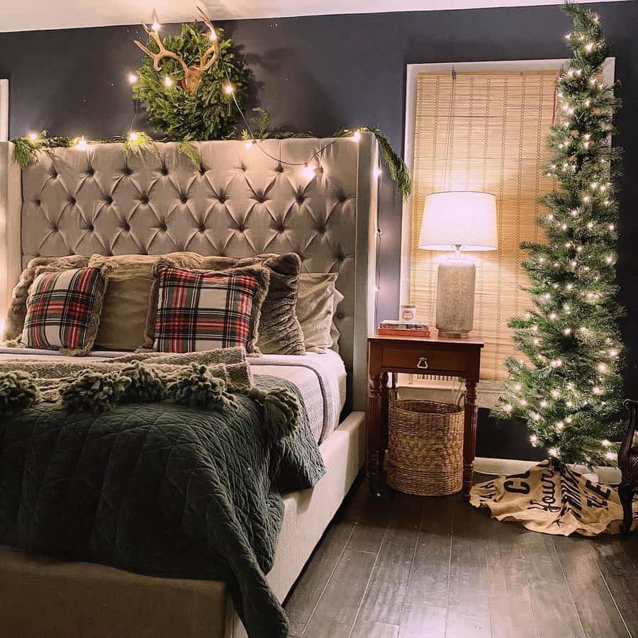 Cozy bedroom with a tufted headboard, plaid pillows, and festive lighting; a small lit Christmas tree and lamp on a side table add warmth