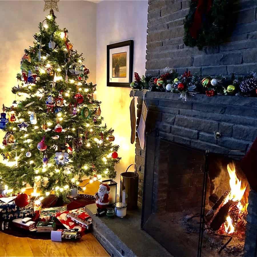 Cozy room with a glowing Christmas tree and a brick fireplace. Gifts are arranged under the tree beside holiday decorations.