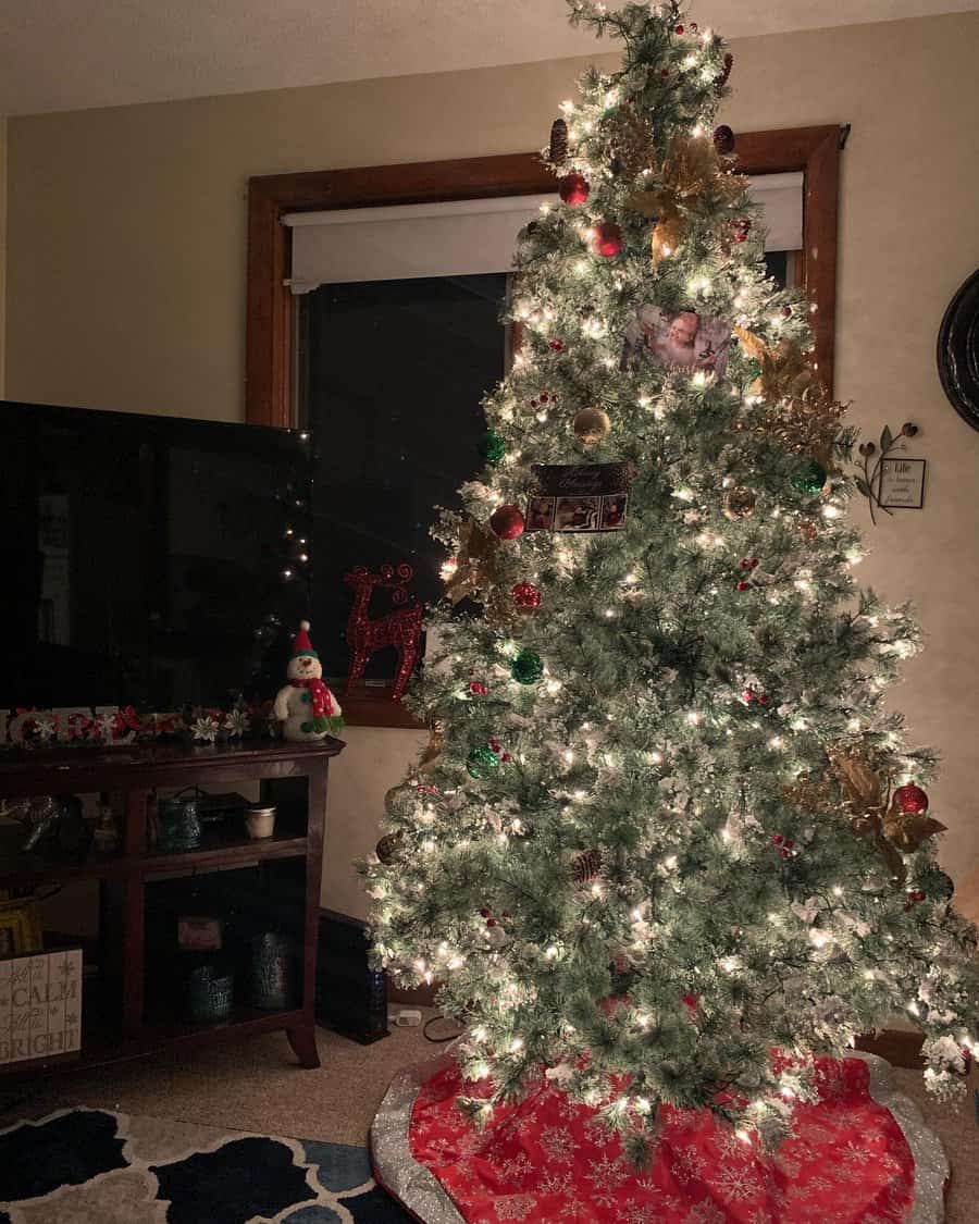 Lit Christmas tree with ornaments and a red tree skirt, next to a TV and snowman decoration in a cozy living room