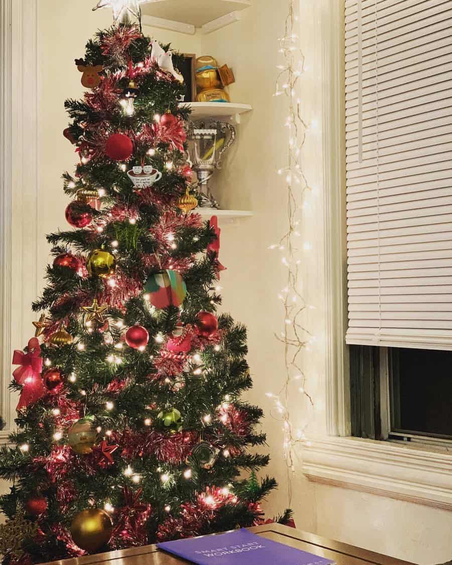 A Christmas tree adorned with red and gold ornaments and lights stands by a window with blinds, next to a shelf with trophies