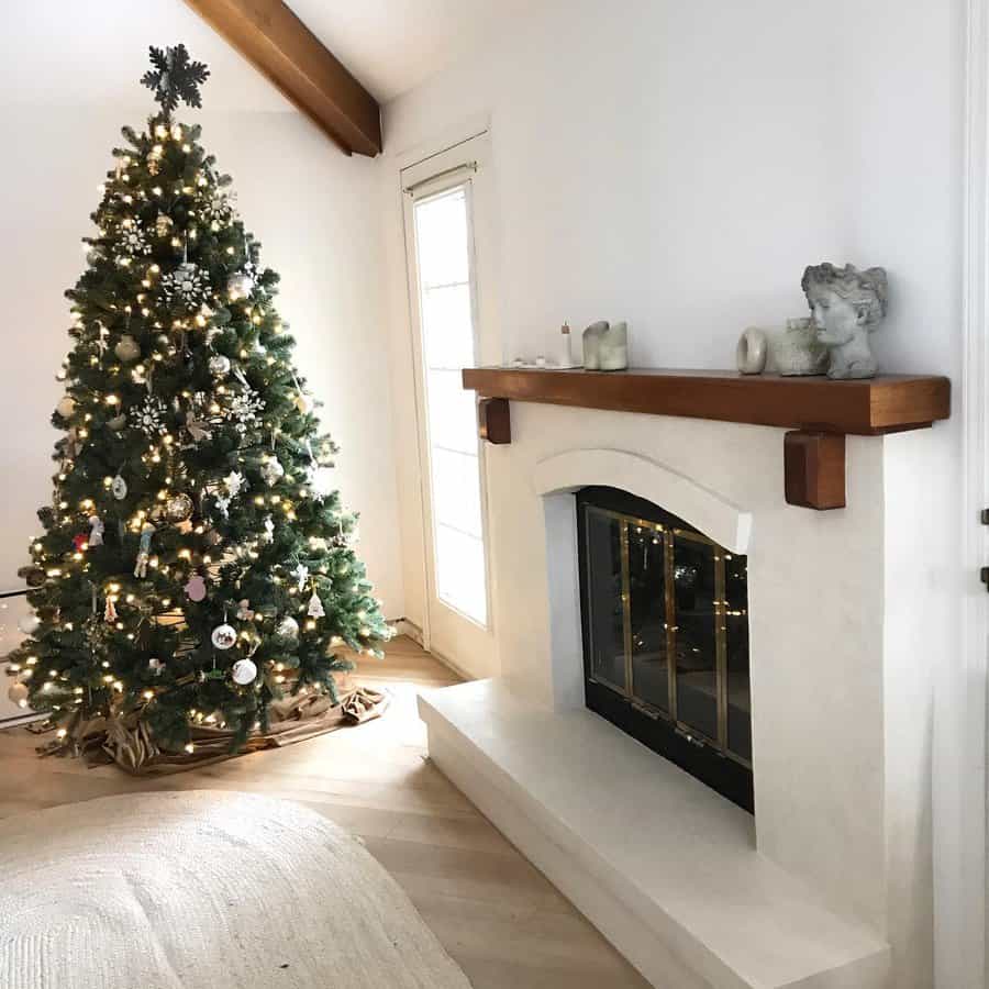 A decorated Christmas tree with lights and ornaments stands next to a white fireplace with a wooden mantel in a cozy living room