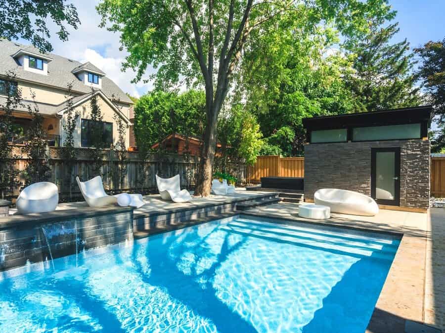 Modern backyard with a clear blue pool, white outdoor furniture, and a small stone structure surrounded by trees and a house