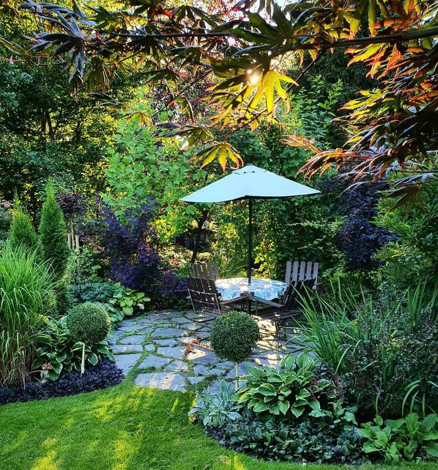 A tranquil garden with a shaded patio, chairs, and a table under a white umbrella, surrounded by lush greenery and sunlight filtering through