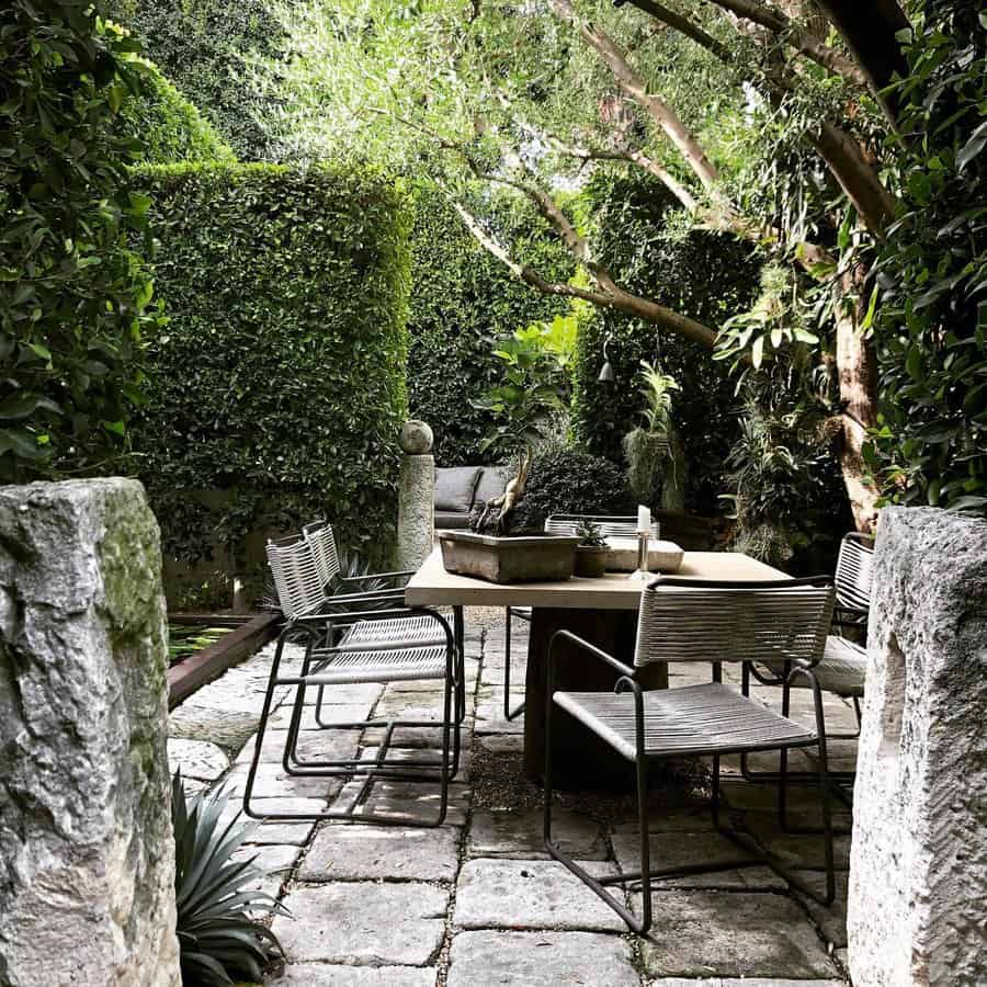 A serene garden patio with a stone path, surrounded by lush greenery, featuring a table and chairs under a large tree