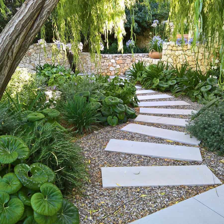 A winding stone path leads through a lush garden with leafy plants, surrounded by a stone wall under a willow tree