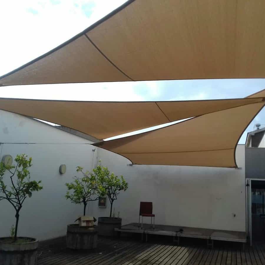 Outdoor courtyard with overlapping beige triangle shade sails, wooden decking, potted trees, and a small stage area