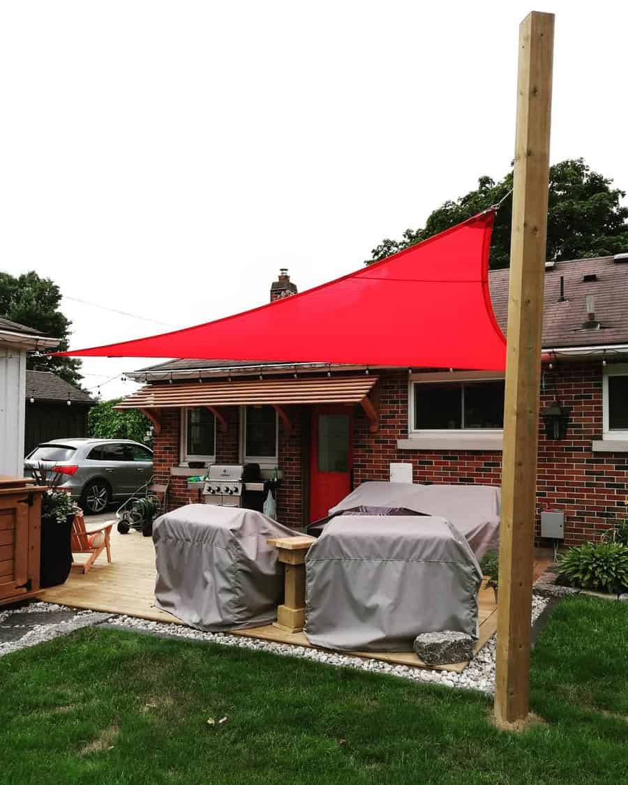 Backyard patio with a bold red triangle shade sail, wooden deck, covered grills, and a brick house with a red door