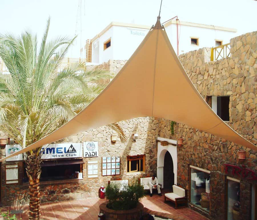 Outdoor courtyard with a large beige triangle shade sail, stone walls, palm trees, and a dive club entrance