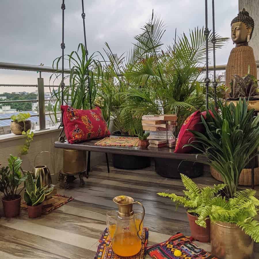 Bohemian-style balcony with lush green plants, a wooden swing, vibrant cushions, a Buddha statue, and a cozy setup for relaxation