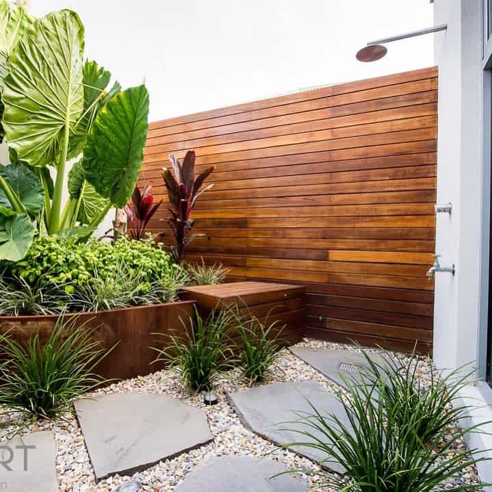 Outdoor shower area with a wooden privacy wall, large tropical plants, stepping stones, and pebble ground cover