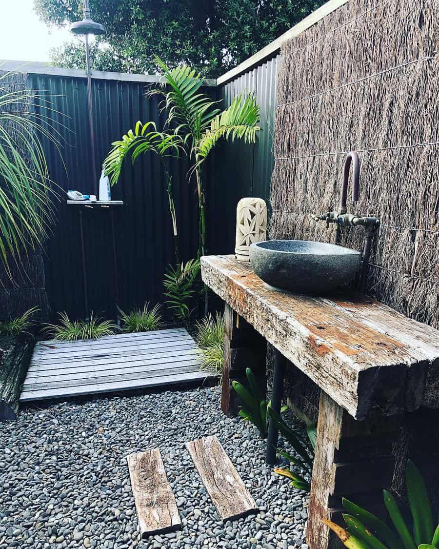 Outdoor bathroom with rustic sink, pebble floor, wooden deck, and lush plants surrounding the space