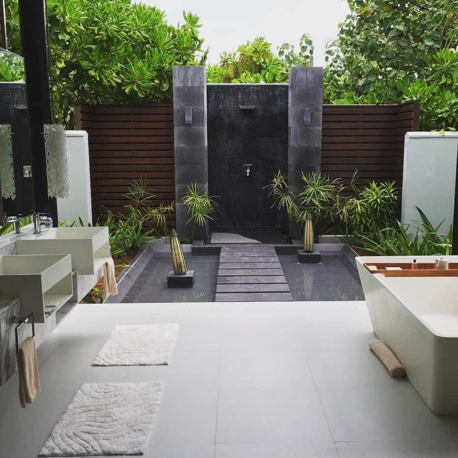 Outdoor bathroom with a modern tub, two sinks, and an open shower surrounded by greenery and wooden accents