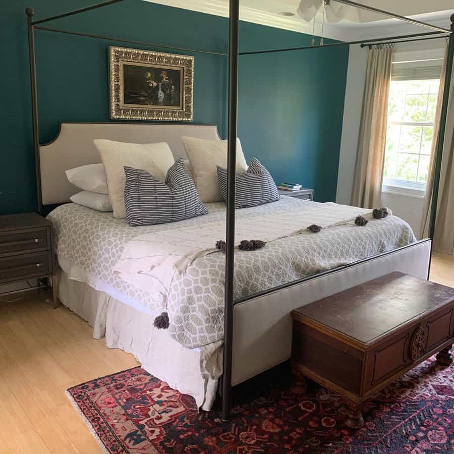 Cozy bedroom with a canopy bed, patterned bedding, decorative pillows, dark green accent wall, a window with curtains, and a wooden chest