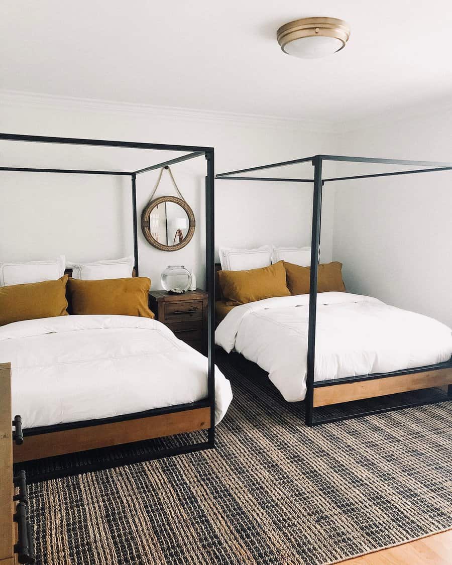 Bedroom with two canopy beds, white bedding, and mustard-colored pillows; a round mirror and a lamp on a wooden nightstand