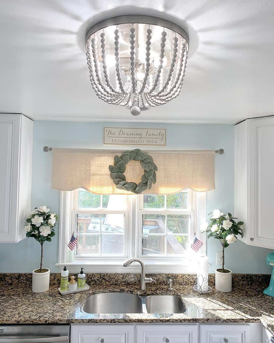 Elegant farmhouse kitchen with a double undermount sink, granite countertops, soft blue walls, a beaded chandelier, and charming window decor