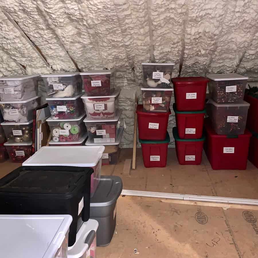 Labeled plastic bins neatly stacked in an unfinished attic with spray foam insulation, organizing holiday decorations and seasonal items.