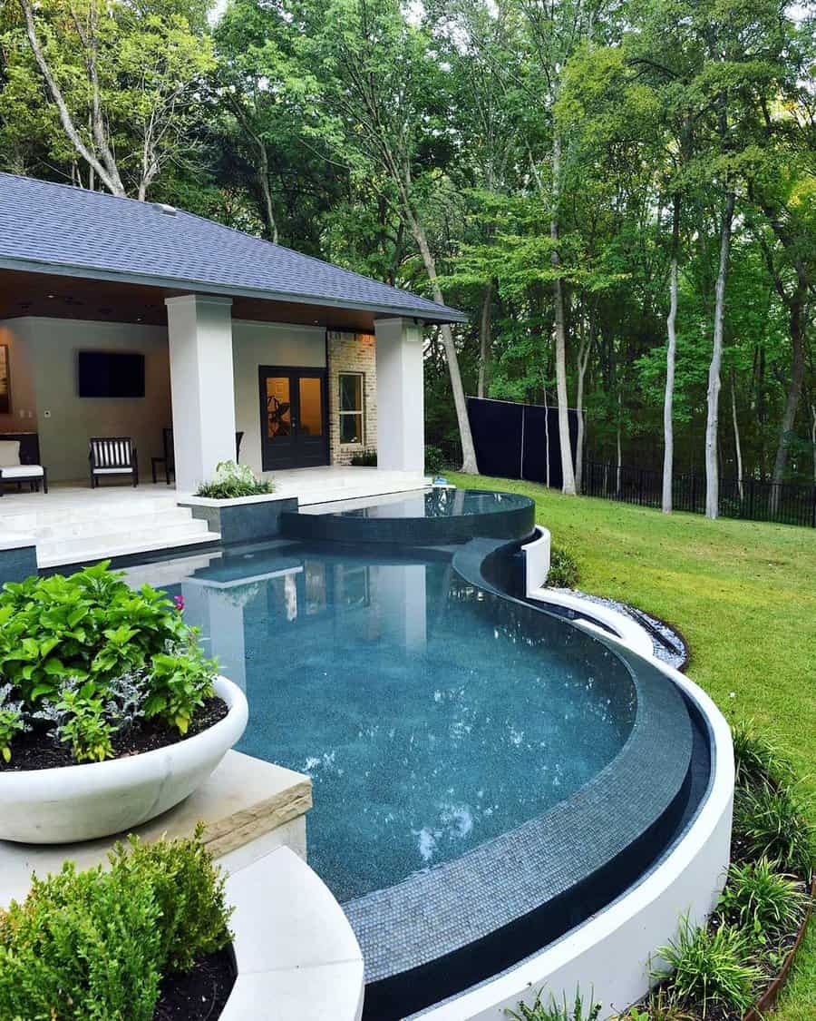 A modern outdoor pool with a curved edge, surrounded by lush greenery and a contemporary house with a patio and seating area