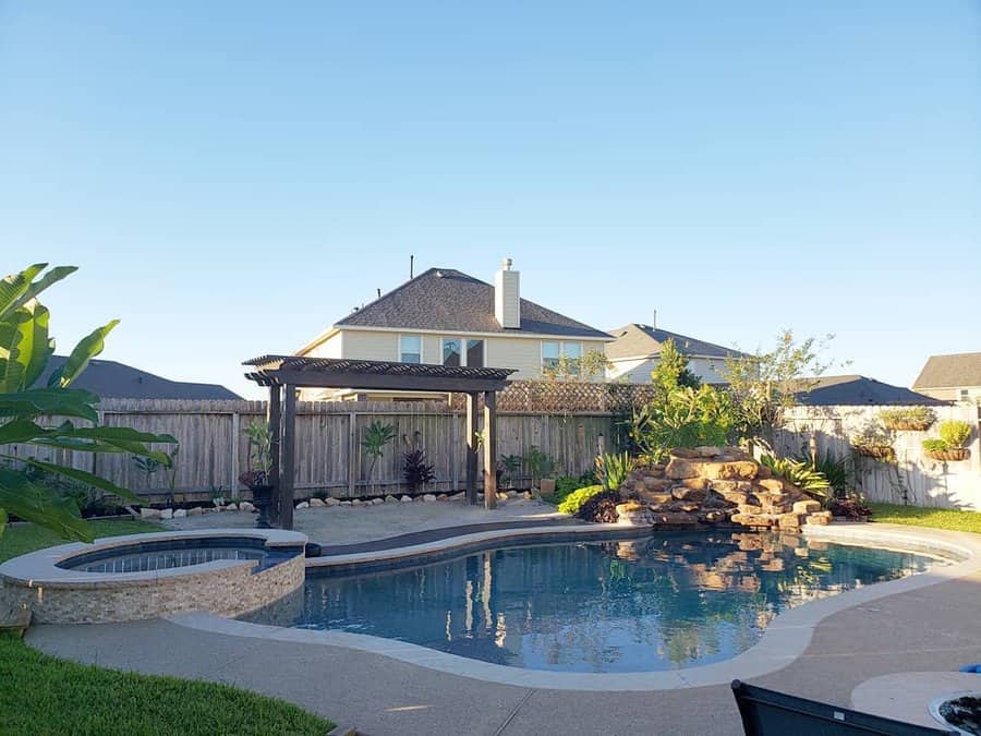 Backyard pool with a waterfall, hot tub, and wooden pergola surrounded by lush greenery under a clear blue sky