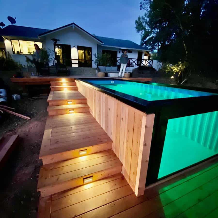 Stylish above-ground pool with wood decking, illuminated steps, and a glass panel for a modern and inviting backyard oasis