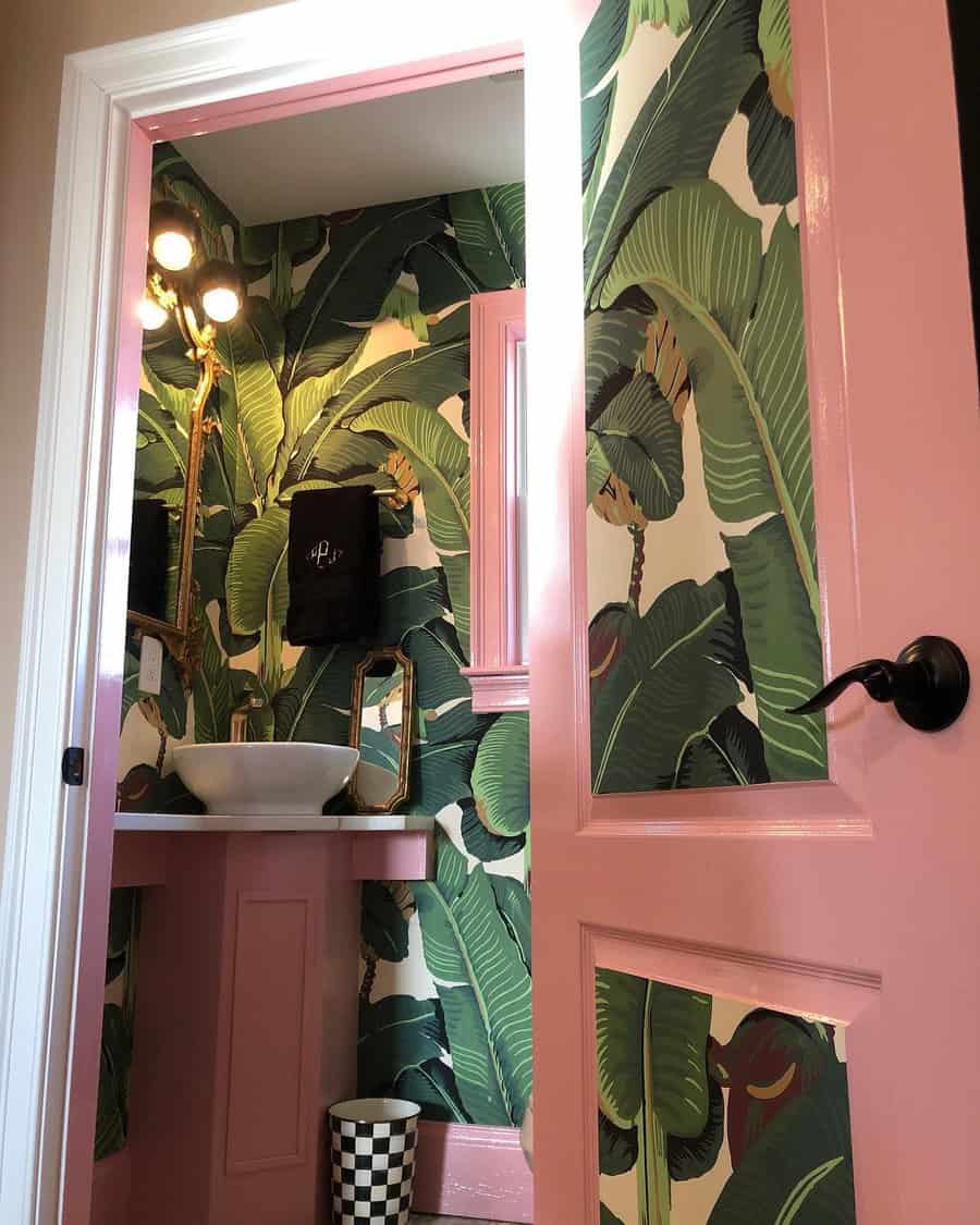 Bathroom with banana leaf wallpaper, pink door and trim, white sink on pink vanity, checkered wastebasket, and wall-mounted light