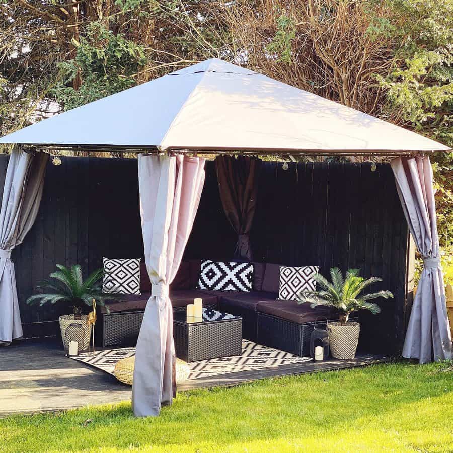 A cozy outdoor gazebo with a seating area, gray curtains, geometric cushions, small palm plants, and lanterns on a grass lawn