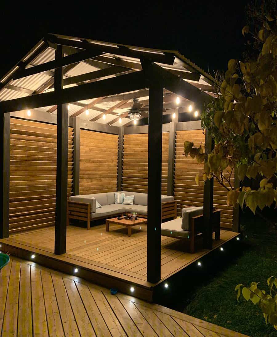 Wooden gazebo at night with cozy seating, string lights, and cushioned chairs on a wooden deck surrounded by darkness