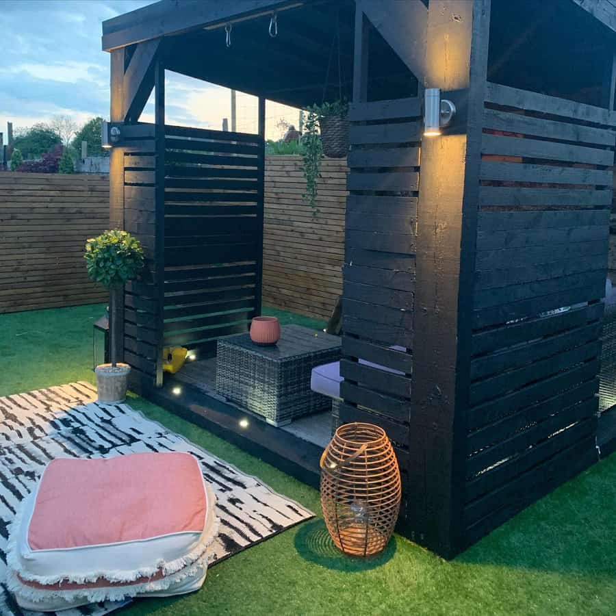 Outdoor wooden pergola with modern seating, cushions, a small table, hanging plants, and ambient lighting on artificial grass