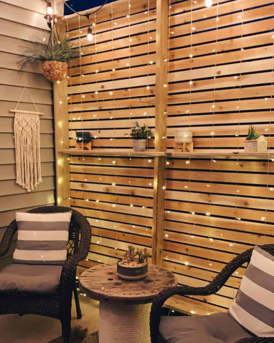 Cozy patio nook with a wooden slat privacy wall, string lights, wicker chairs, striped cushions, and rustic decor for a warm ambiance